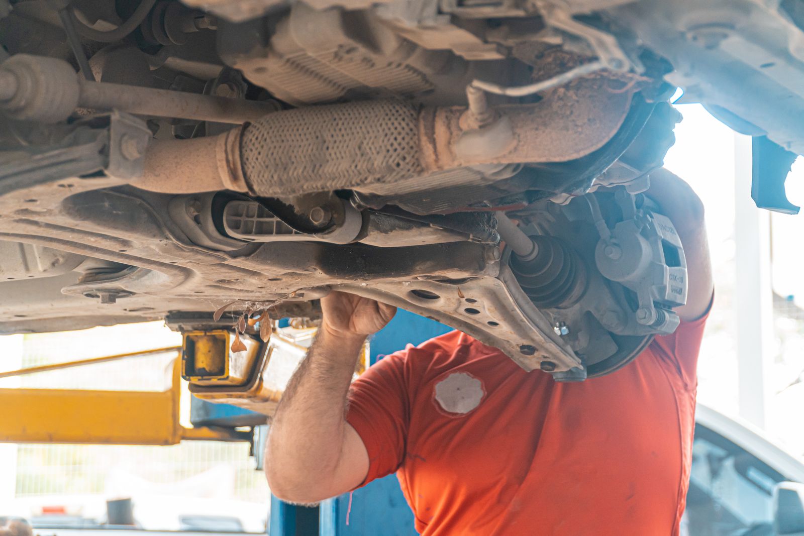 A mechanic in an orange t-shirt reaches up to the front suspension of a lifted car, with a driveshaft rubber CV boot and a brake caliper in the foreground.