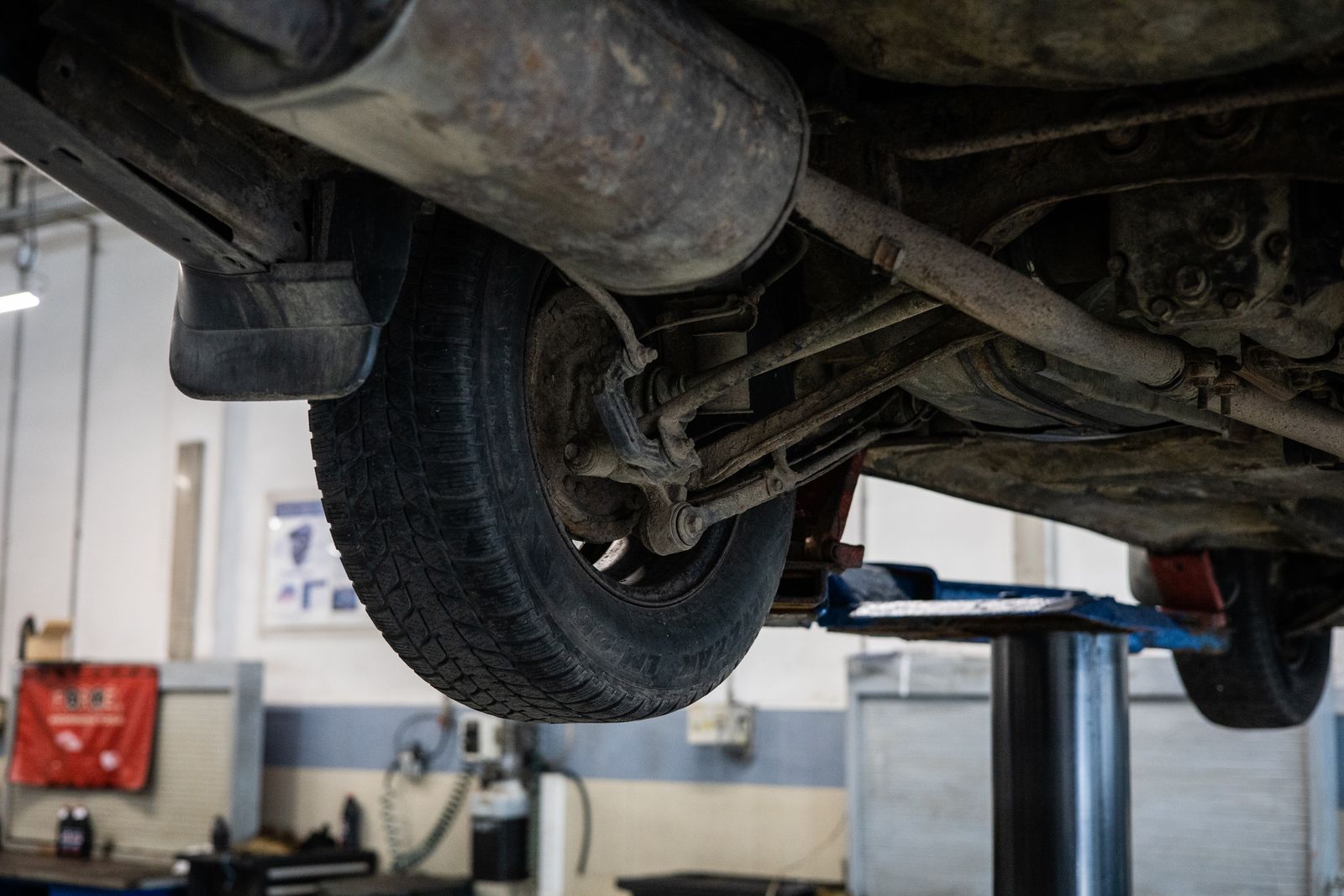A car raised on a workshop lift seen from below, showing the underside, exhaust and rear suspension linkage, with a blurred workshop in the background.