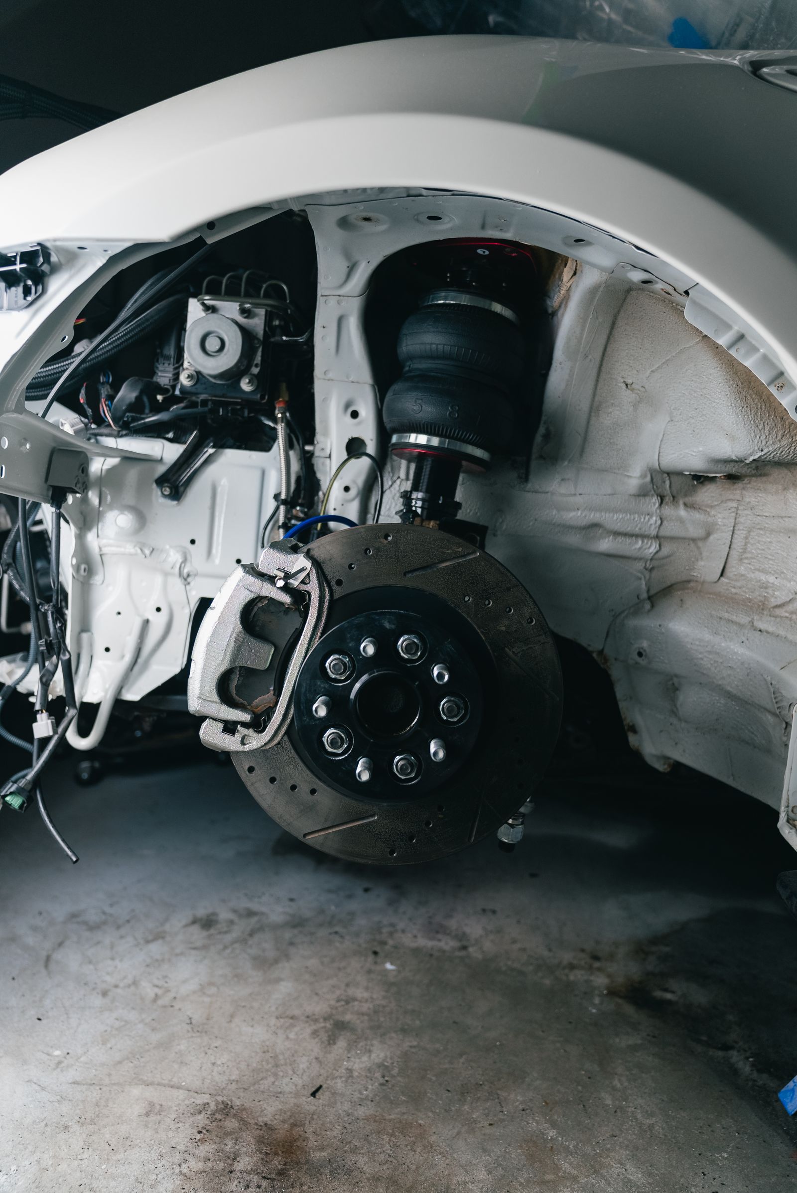 Close-up of a vehicle's front wheel arch with the wheel removed, showing a drilled brake disc, caliper, wheel studs and a black air-spring suspension unit above.